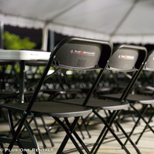 A photo of several black folding chairs with stickers that read, "Plus One Rentals," on the back of the chairs, set up at white folding tables, under a large frame tent.